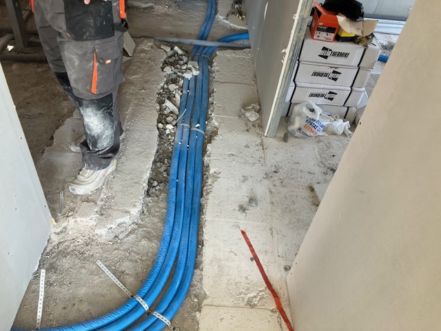 Blue water and sewage lines laid into a floor trench as services are routed between first-floor rooms.